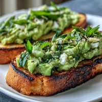 Vibrant fava bean and mint bruschetta on grilled sourdough, topped with creamy ricotta and fresh herbs.  